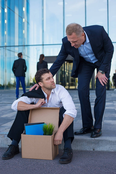 Fired business man sitting frustrated and upset on the street near office building with box of his belongings. He lost work. Other businessman comforts and encourages him - Photo, Image