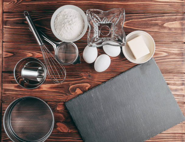 Ingredients in the dishes on a wooden brown table, where as well lie the whisk is full for flour, three chicken eggs and a slate board. View from above. Place under the text - Foto, Imagen