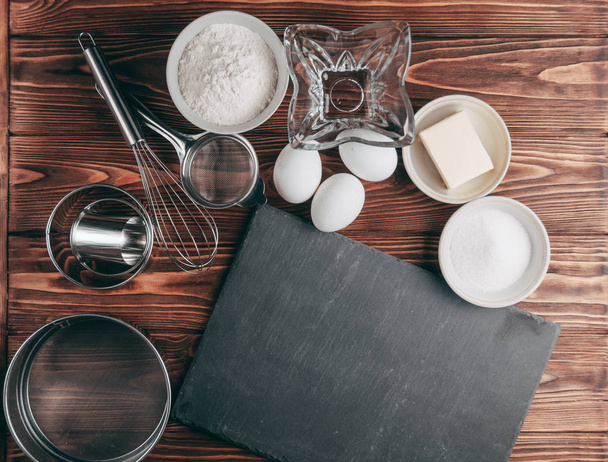 Ingredients in the dishes on a wooden brown table, where as well lie the whisk is full for flour, three chicken eggs and a slate board. View from above. Place under the text - Fotografie, Obrázek