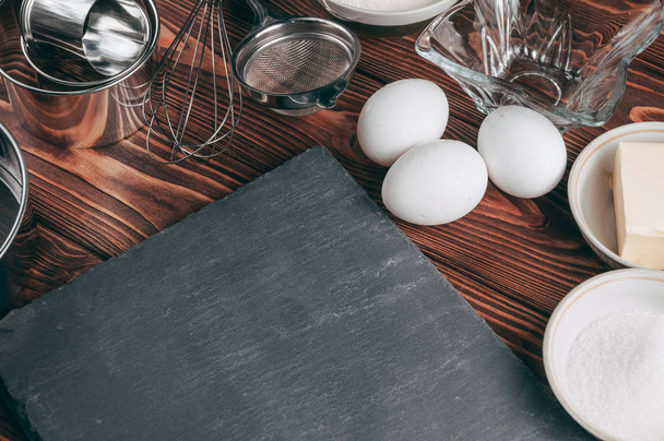 Ingredients in the dishes on a wooden brown table, where as well lie the whisk is full for flour, three chicken eggs and a slate board. View from above. Place under the text - Foto, imagen