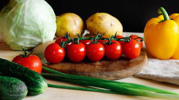 Verduras frescas en la tabla de cortar en la cocina. Tomates cherry. pepino, pimiento amarillo, papa, repollo. Mesa de madera, fondo negro
. - Foto, Imagen