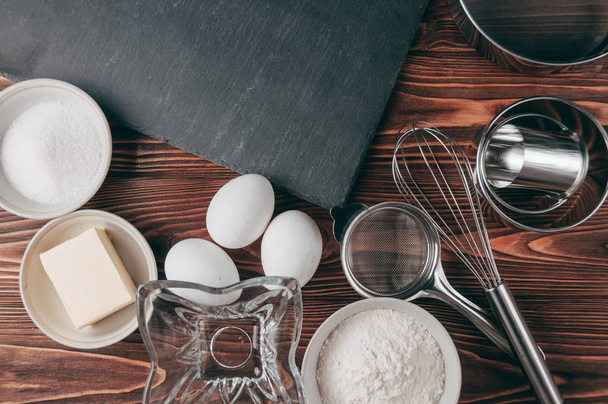 Ingredientes nos pratos em uma mesa marrom de madeira, onde também se encontra o batedor está cheio de farinha, três ovos de galinha e uma tábua de ardósia. Vista de cima. Colocar sob o texto
 - Foto, Imagem