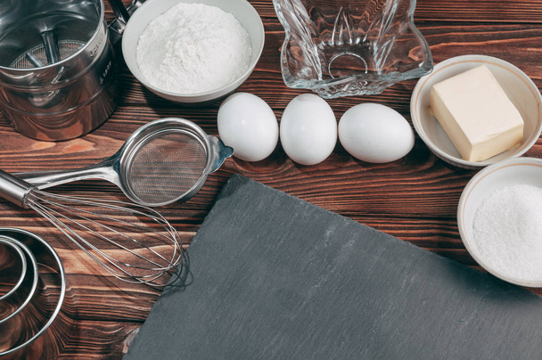 Ingredienti nei piatti su un tavolo di legno marrone, dove si trova anche la frusta è piena di farina, tre uova di pollo e una lavagna. Vista dall'alto. Inserire sotto il testo
 - Foto, immagini