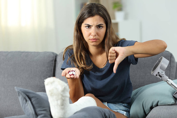 Angry disabled woman holding pills with thumbs down sitting on a couch in the living room at home - Foto, Imagem