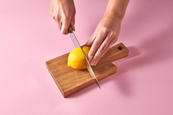 Citrus fruits on cutting board. Female hands cutting a yellow ripe lemon on half on a wooden board on a pink background. Concept vegetarian food. - Фото, изображение