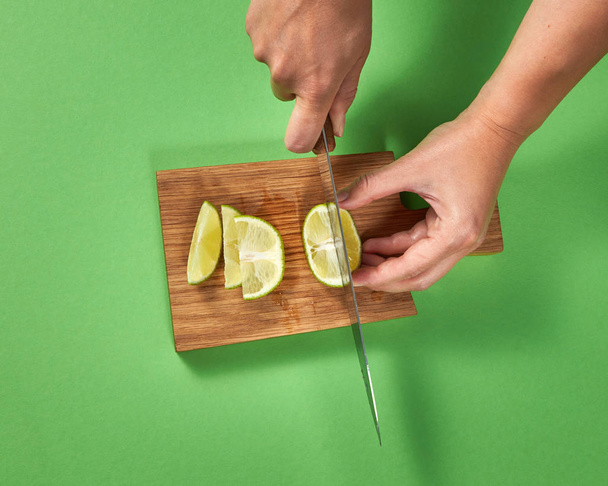 Top view of woman cutting a half of organic ripe lime with slices on a brown board on a green background. A sharp knife in her hands. - Foto, immagini