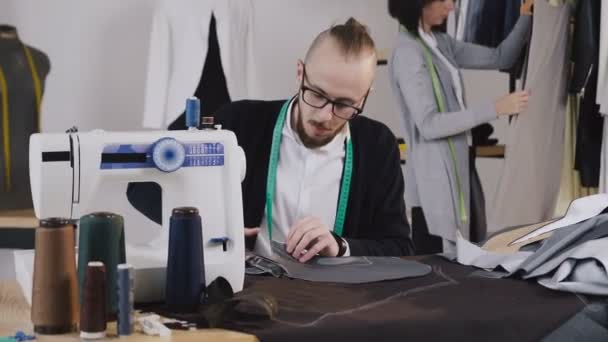 Handsome tailor sits on the workplace at studio and cutting gray fabric using large scissors as he follows the chalk markings of the pattern using pattern. In the background, the dressmaker looks at - Кадры, видео