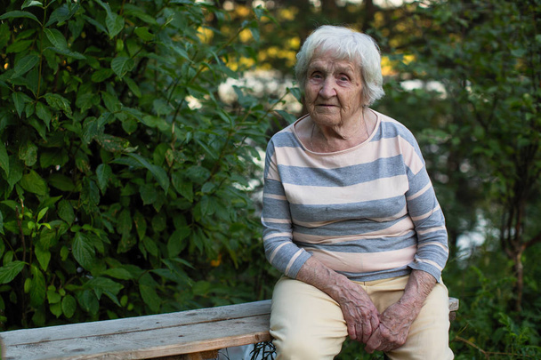 Μια ηλικιωμένη γυναίκα που κάθεται σε ένα ξύλινο παγκάκι στο πάρκο. - Φωτογραφία, εικόνα