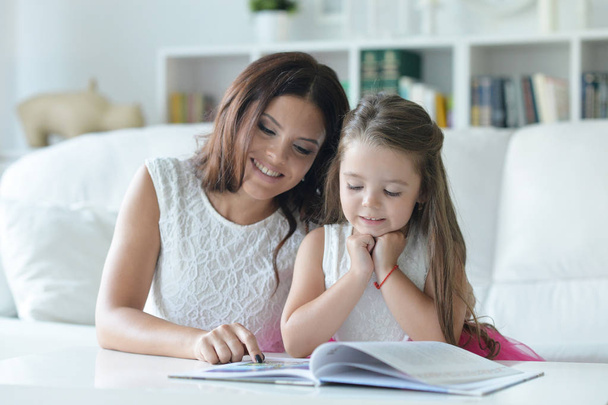 маленькая милая девочка читает книгу с мамой за столом дома
 - Фото, изображение