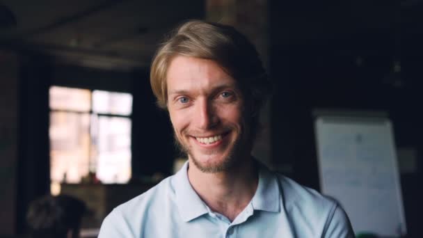 Close-up slow motion portrait of happy young man entrepreneur standing in workplace, looking at camera and smiling. Modern office with people is in background. - Materiał filmowy, wideo