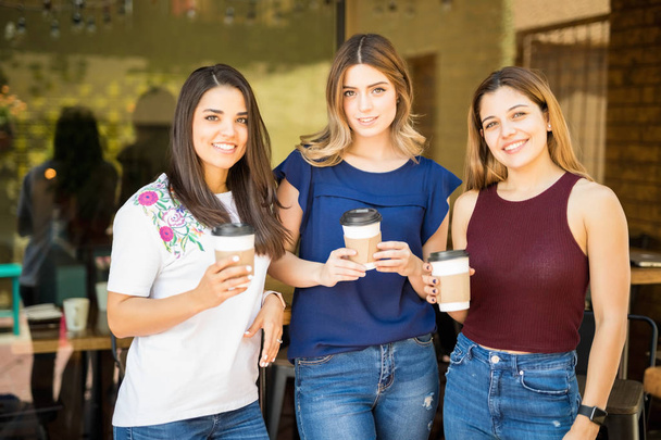 Retrato de três amigas do sexo feminino em pé do lado de fora de um restaurante com xícaras de café e fazendo contato visual
 - Foto, Imagem