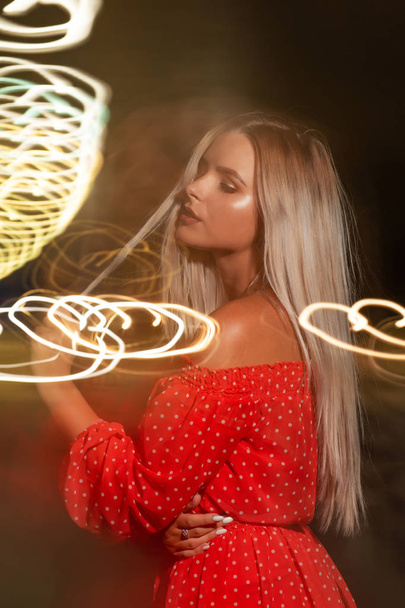 Adorable young blonde woman wearing red dress posing with blurres night light - Фото, изображение
