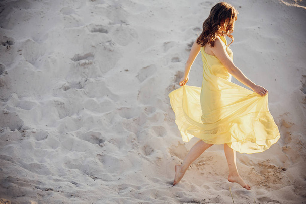 longitud completa de hermosa chica en vestido amarillo que se divierten caminando en la playa de arena en la luz del atardecer cerca del lago. concepto de vacaciones de verano
 - Foto, Imagen