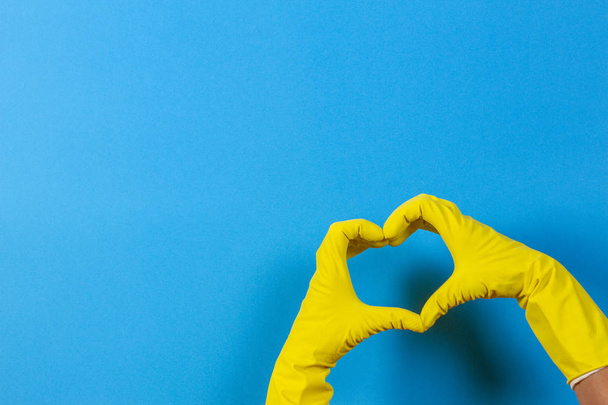 Hands in yellow rubber gloves making heart shape with fingers, on blue background - Fotoğraf, Görsel