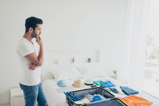 Time to plan a trip! Young brunet man packing suitcase in a bright bedroom, looks at things thoughtful touch hand to chin - Photo, image