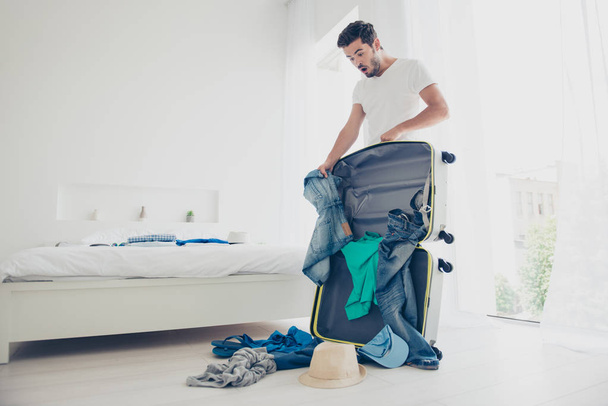 Handsome young bearded shocked confused brunet man wearing white t-shirt, packing luggage to a trip, journey, room in mess - 写真・画像