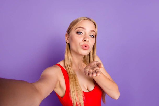 Self portrait of attractive cheerful playful blonde funny gorgeous girl wearing red swim suit sending you air kiss with pouted lips. Isolated over violet pastel background - Foto, afbeelding