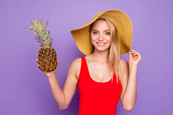 Close up portrait of beautiful and kind girl holds a pineapple in her hand and looks into the camera holding her hat by hand isolated on violet background - Φωτογραφία, εικόνα