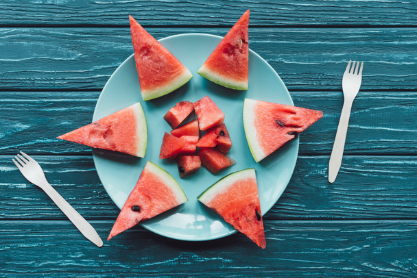 flat lay with arranged watermelon pieces on plate and plastic forks on blue wooden surface - Foto, Imagen
