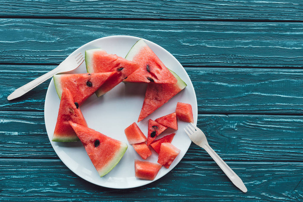 flat lay with arranged watermelon pieces on plate and plastic forks on blue wooden surface - Fotó, kép