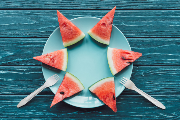 vlakke leggen met watermeloen gearrangeerde stukken op plaat en plastic vorken op blauwe houten oppervlak - Foto, afbeelding