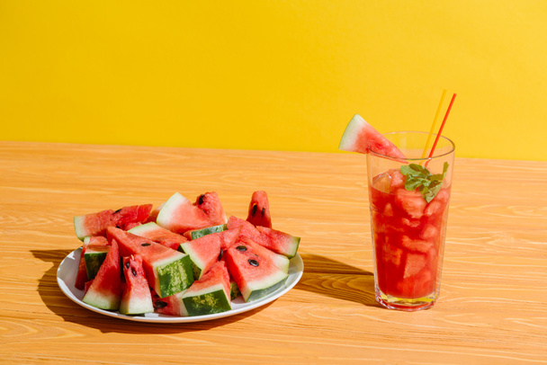 close up view of fresh watermelon pieces and watermelon drink in glass on wooden surface on yellow background - Foto, imagen