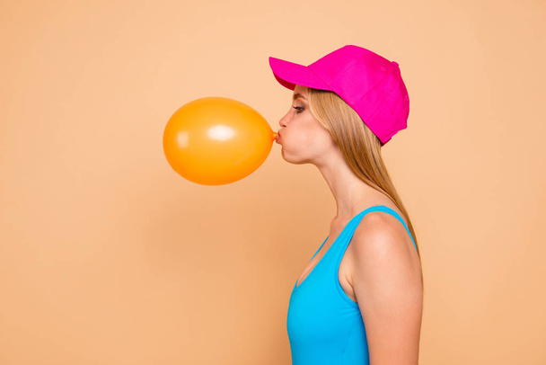 Portrait of young attractive nice cute straight-haired blonde funky girl wearing casual, bright pink red cap, blowing yellow air baloon. Side profile view. Isolated over beige background - Photo, Image