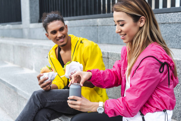 sonriente asiático mujer apertura soda beber mientras su novio holding hamburguesas en urbano calle
 - Foto, imagen