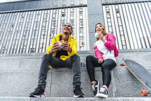 low angle view of young smiling multicultural couple eating burgers near skateboard at city street - Valokuva, kuva