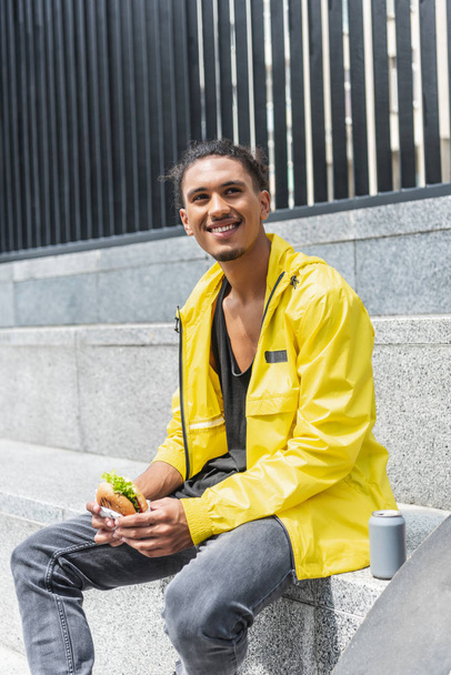 smiling mixed race man having lunch with burger and soda at city street - Φωτογραφία, εικόνα