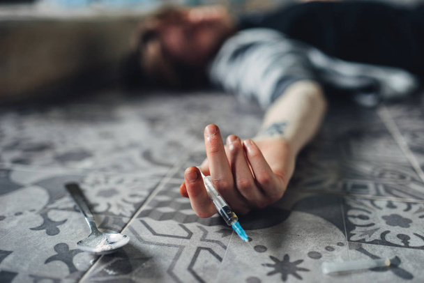 Drug addict lies on the floor, hand with syringe and spoon for dose preparing on the table. Addiction concept, addicted people - Photo, Image