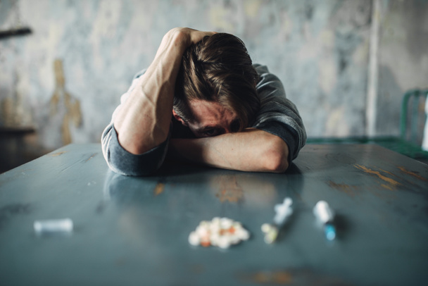 Drogato di sesso maschile seduto al tavolo con droghe e siringhe, interno della sala grunge sullo sfondo. Concetto di tossicodipendenza, tossicodipendenti
 - Foto, immagini