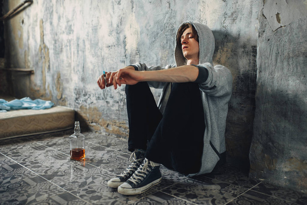 Junkie with syringe in hand sitting on the floor after dose, bottle of alcohol is near. Drug addiction concept, narcotic addicted people - Fotografie, Obrázek