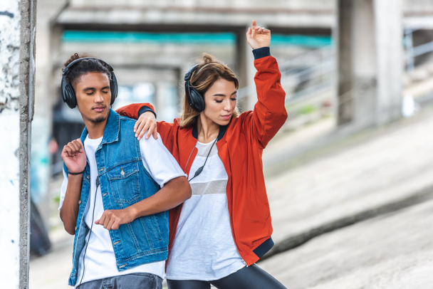 stylish multicultural couple listening music with headphones and dancing at city street  - Foto, Bild