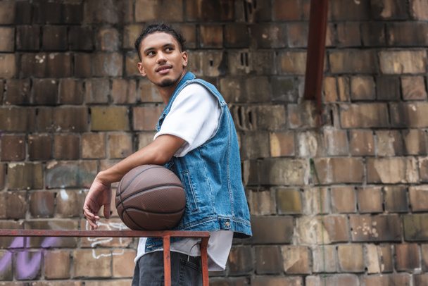 stylish mixed race man standing with basketball ball and looking away at street - Foto, imagen