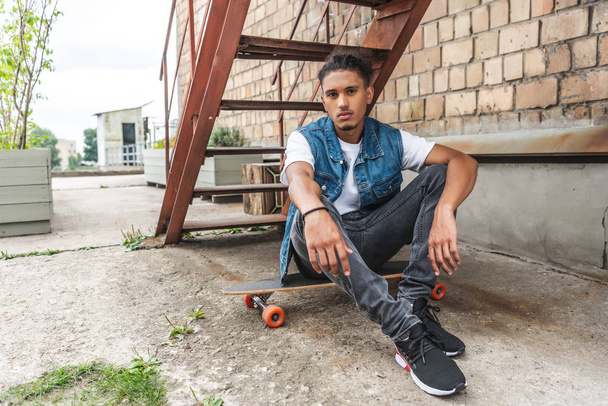 young stylish mixed race man sitting on skateboard and looking at camera at urban street - Фото, изображение