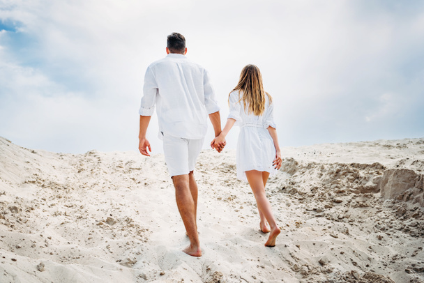 Rückansicht eines jungen Paares in weißer Kleidung, das barfuß am Sand entlang geht - Foto, Bild