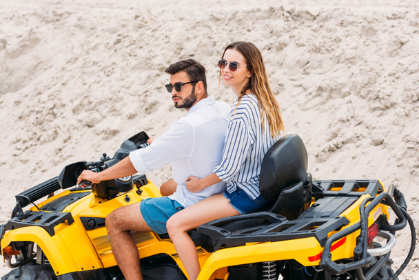 beautiful young couple in sunglasses riding all-terrain vehicle in desert - Photo, Image
