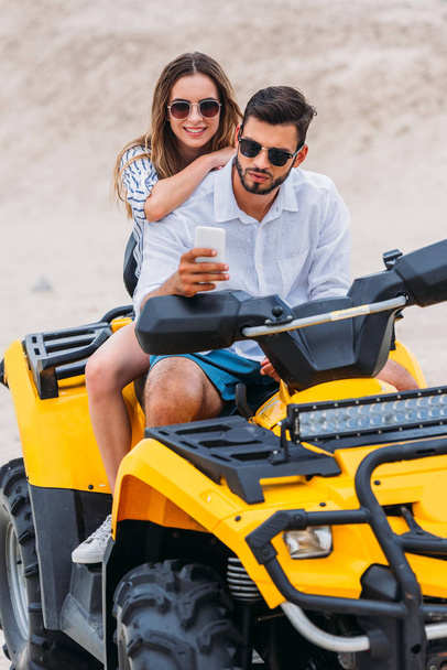beautiful young couple taking selfie while sitting on ATV in desert - Photo, Image