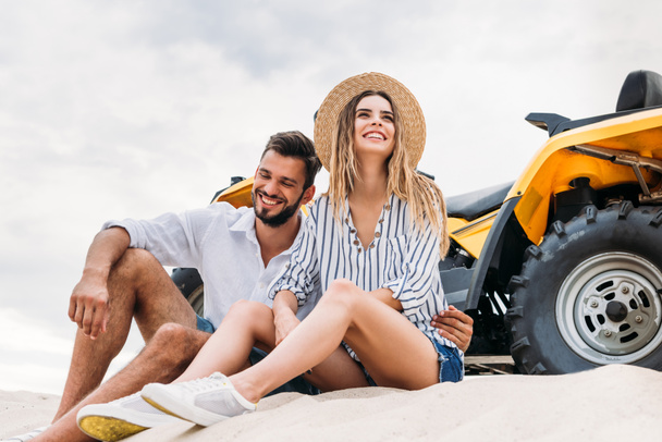 sonriente joven pareja sentado cerca de ATV en duna de arena
 - Foto, Imagen