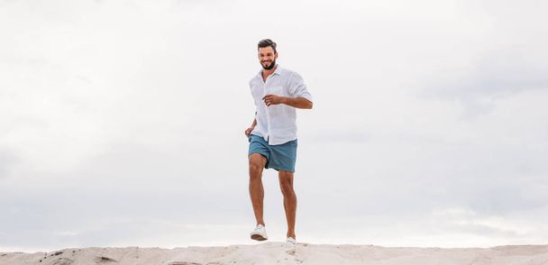handsome young man running on sand dune in front of cloudy sky - Photo, Image