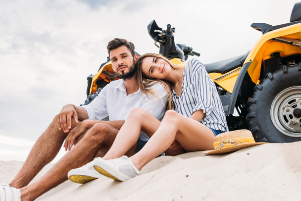 bottom view of beautiful young couple sitting near ATV on sandy dune and looking at camera - Foto, Bild