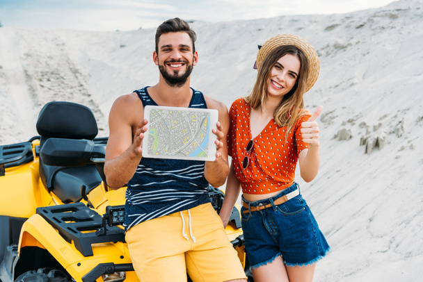 happy young couple with atv showing digital tablet with map on screen and thumb up at desert - Фото, изображение