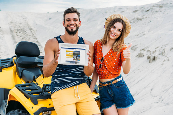 beautiful young couple with atv showing digital tablet with booking website on screen and thumb up at desert - Foto, imagen