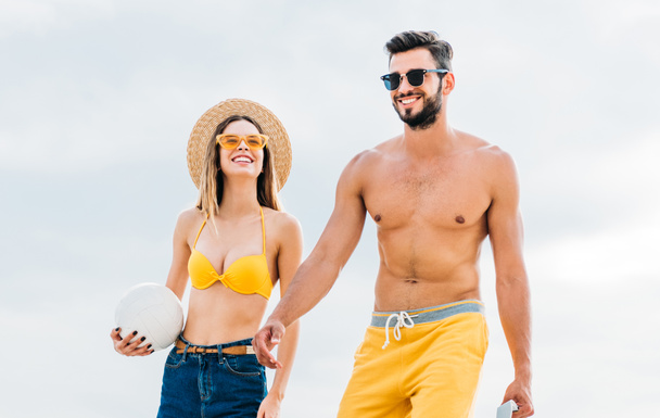 beautiful young couple in beach clothes with volleyball ball in front of cloudy sky - Фото, изображение