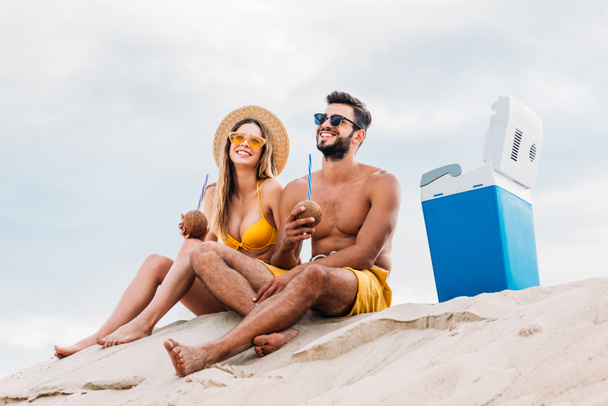 sonriente pareja joven con cocteles de coco relajándose en la arena frente al cielo nublado
 - Foto, Imagen