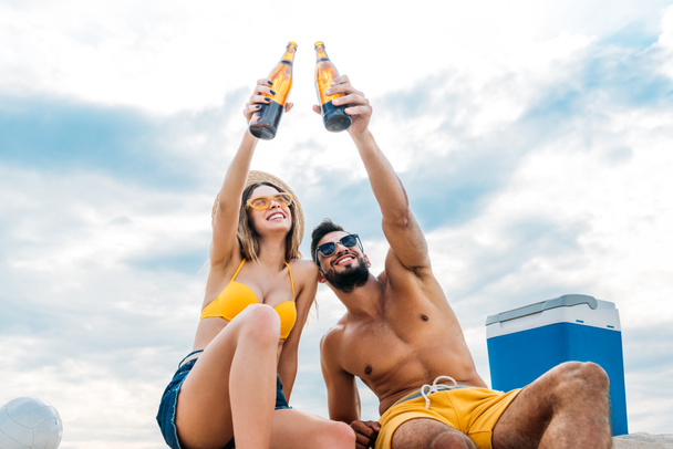 vue du bas de beau jeune couple soulevant des bouteilles de bière devant le ciel nuageux tout en étant assis sur le sable
 - Photo, image