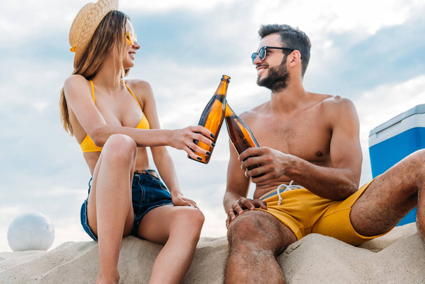 bella giovane coppia clinking con bottiglie di birra mentre seduto sulla sabbia
 - Foto, immagini