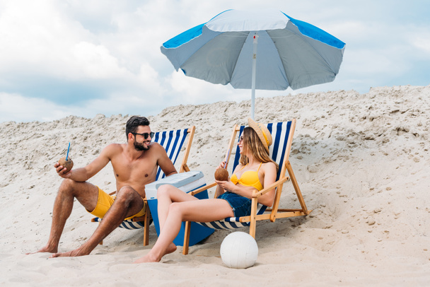 beautiful young couple with coconut cocktails relaxing in sun loungers on sandy beach - Photo, Image