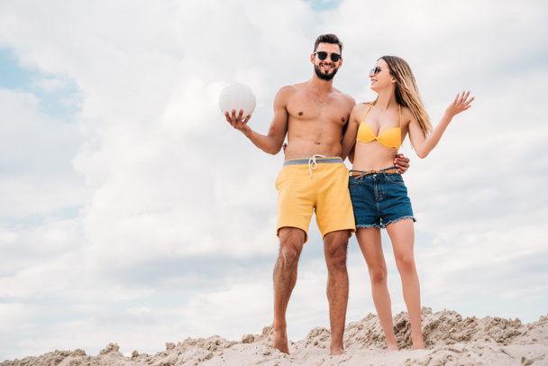 beautiful young couple with volleyball ball standing on sandy dune in front of cloudy sky - Photo, Image